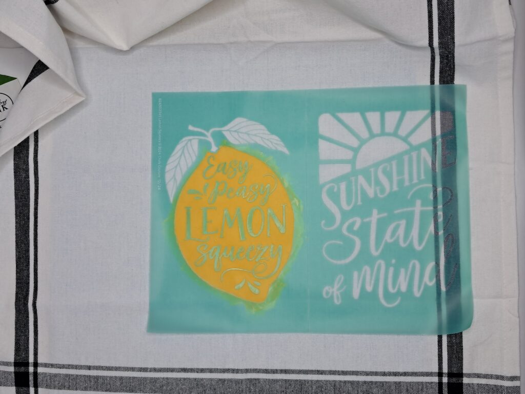 Adhesive silk screen mesh stencil is placed on top of the tea towel. Yellow permanent ink has been added to the stencil.