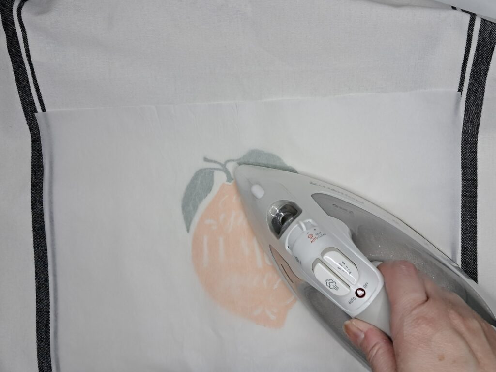 Stenciled tea towel is protected by a sheet of parchment paper while being heat set with an iron.